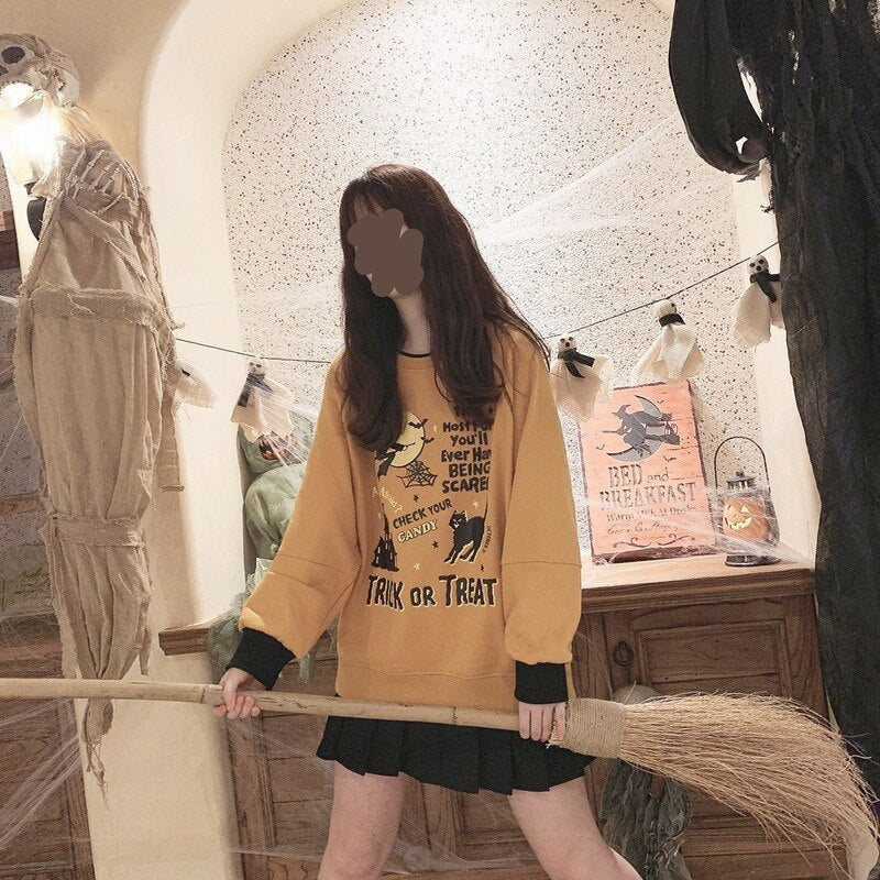 Model wearing the orange Halloween graphic sweatshirt and black skirt while holding a broom horizontally in a decorated room.