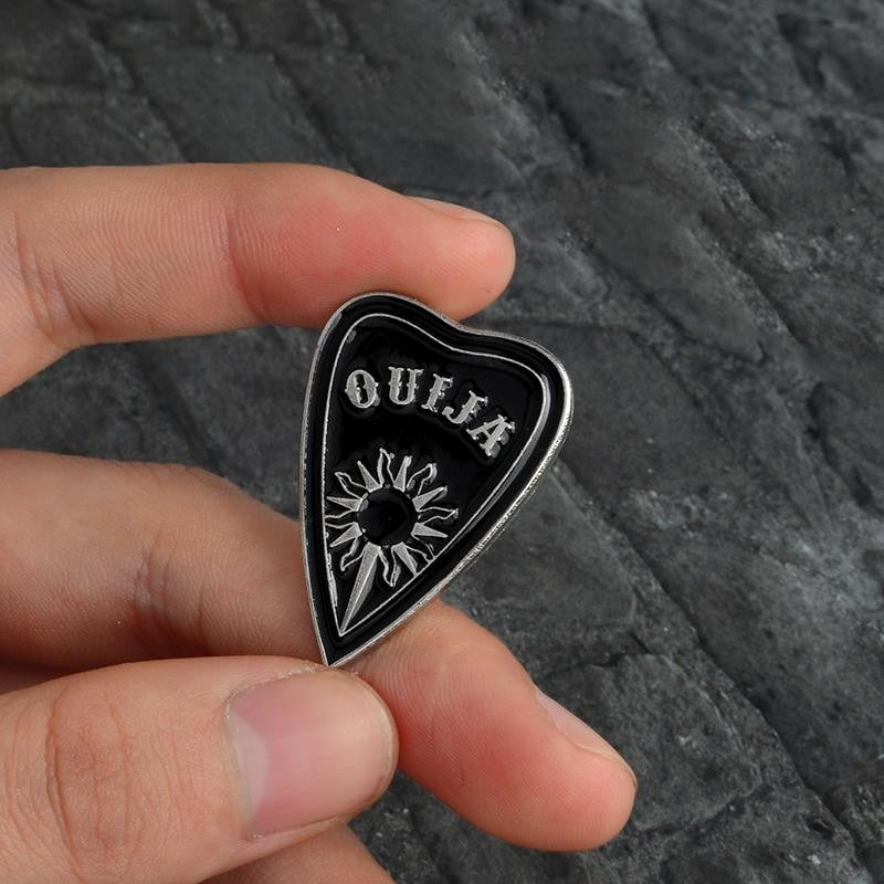 Black Ouija planchette pin held between fingers, with its silver-tone sun, round opening and triangular outline visible.