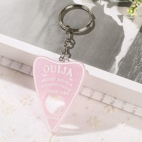Pale pink glitter planchette keychain with white lettering and a white outline, displayed on white lace with the ring and chain resting above it.