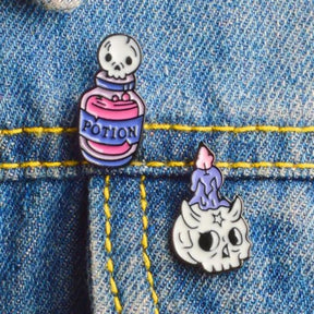 Potion-bottle and skull-candle pins attached beside a blue denim seam, showing their pink, purple and white artwork outlined in black.