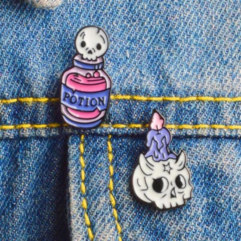 Potion-bottle and skull-candle pins attached beside a blue denim seam, showing their pink, purple and white artwork outlined in black.