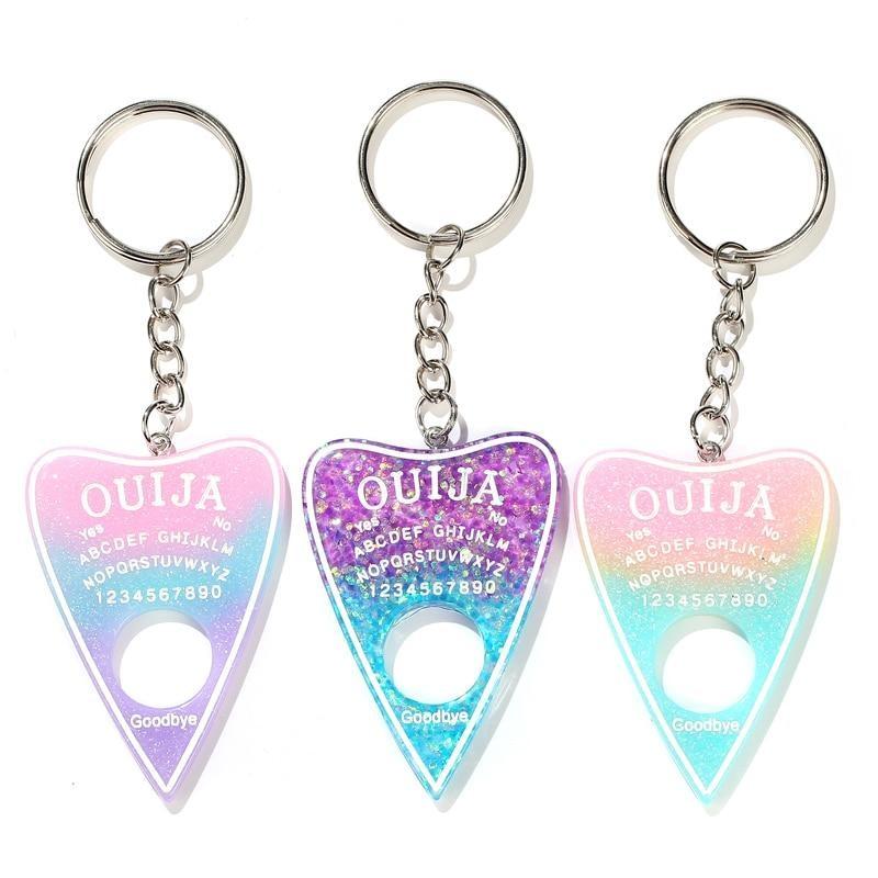 Three planchette resin keychains arranged on white: pink-blue-purple, purple-blue glitter and pink-yellow-aqua gradients. Each has white spirit-board lettering, a round cutout and a silver-tone keyring.