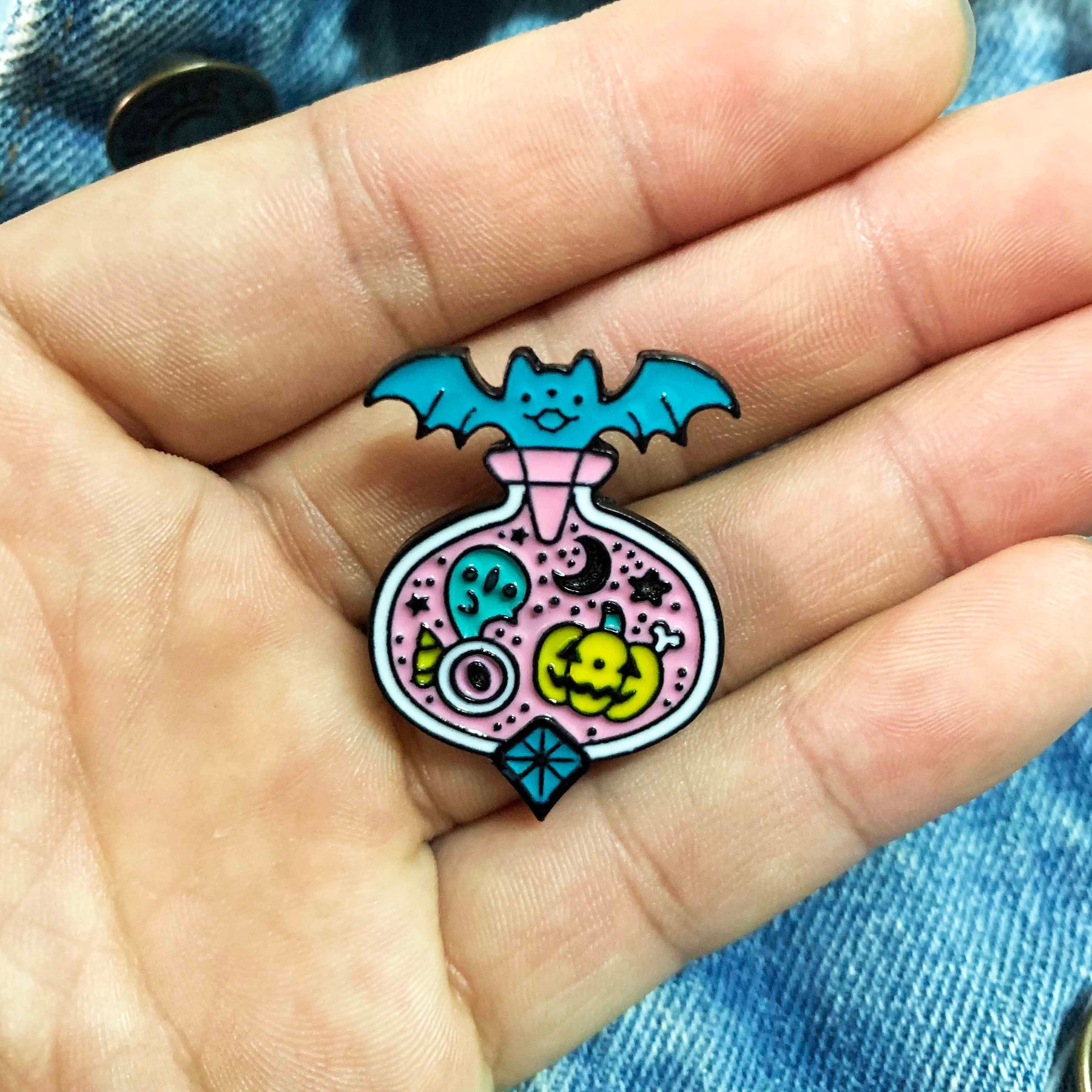 Small lavender and turquoise Halloween potion pin resting in an open palm above blue denim, showing its scale against the hand.