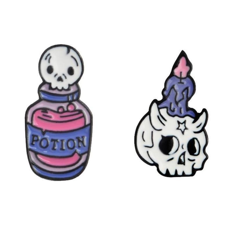 Two pastel goth enamel pins on white: a pink potion bottle with a skull stopper and a horned white skull holding a purple candle.