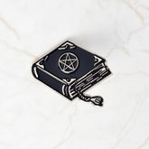 Black spellbook enamel pin on pale marble, with a silver-tone pentacle, decorative corner marks, outlined page edges and a curling tasselled bookmark.