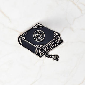 Black spellbook enamel pin on pale marble, with a silver-tone pentacle, decorative corner marks, outlined page edges and a curling tasselled bookmark.