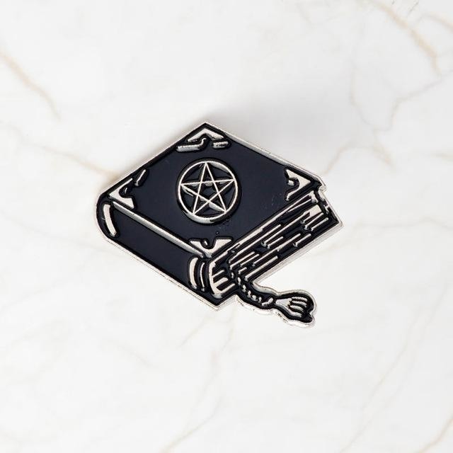 Black spellbook enamel pin on pale marble, with a silver-tone pentacle, decorative corner marks, outlined page edges and a curling tasselled bookmark.