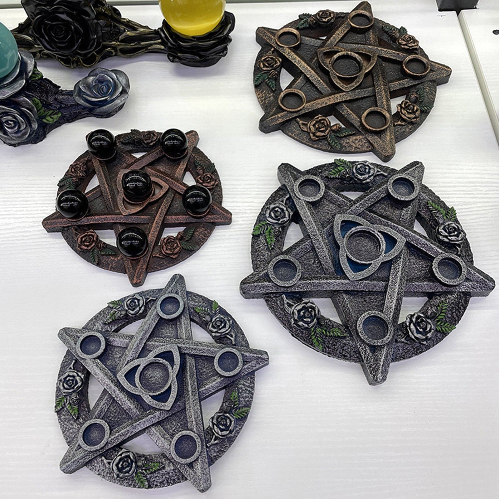 Several rose-and-triquetra candle holders in metallic-tone finishes arranged on a white display surface, including extra display variations.