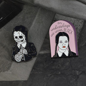 Two gothic portrait pin designs arranged on dark gray textured fabric, with the pink arched slogan pin beside the skeletal figure.