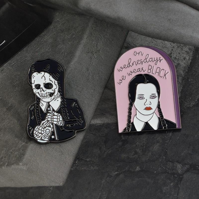 Two gothic portrait pin designs arranged on dark gray textured fabric, with the pink arched slogan pin beside the skeletal figure.