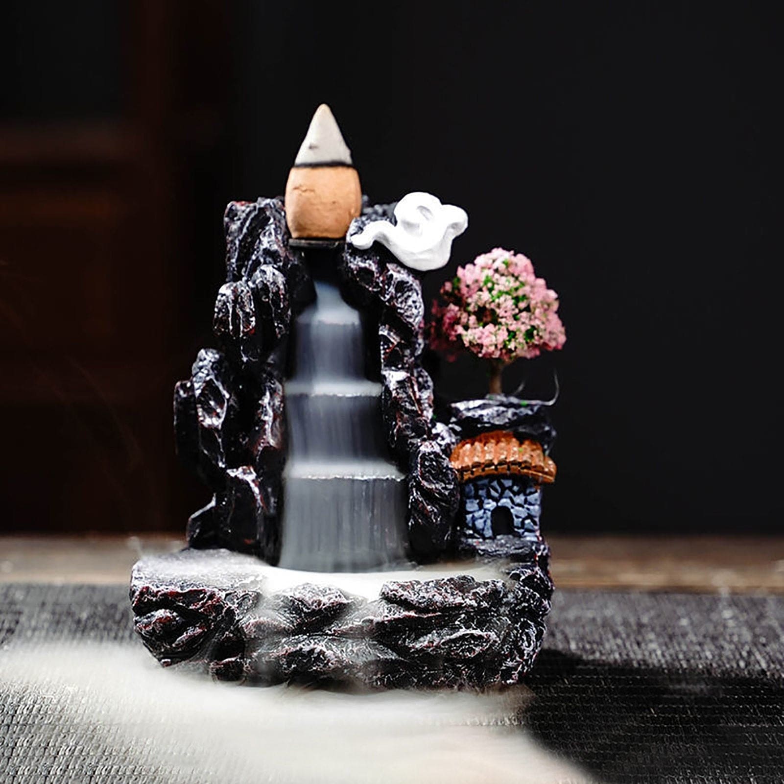 The dark village burner against a shadowed background, with bright white smoke descending three steps beside the pink blossom tree and tiled roof.
