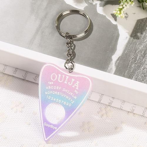 Pastel pink, blue and purple gradient planchette keychain with white alphabet and number details, displayed on lace at the edge of an open book.