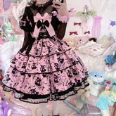 Pink tiered Halloween dress with oversized bows, black front lacing and a print of pointed-hat figures, bats, crosses and spiderwebs, worn over a sheer black blouse.