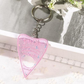 Translucent pink planchette resin keychain with iridescent flecks and pink lettering, displayed on white lace with its silver-tone keyring above the charm.