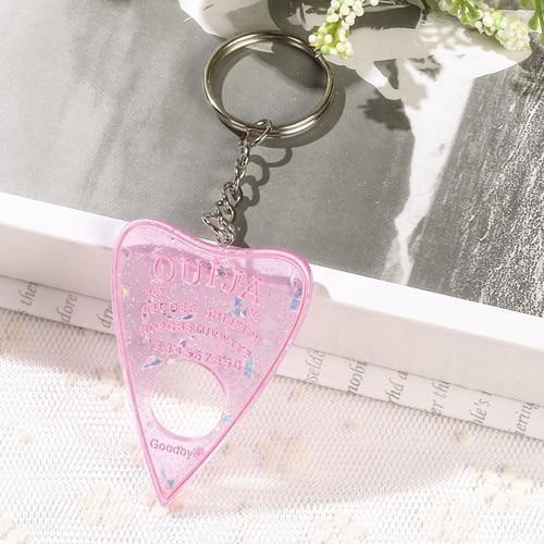 Translucent pink planchette resin keychain with iridescent flecks and pink lettering, displayed on white lace with its silver-tone keyring above the charm.