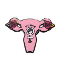 Pink uterus-shaped enamel pin with Woman Up lettering, roses at both sides and a raised fist inside the female symbol at the centre.