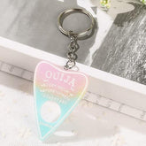 Planchette keychain fading from pastel pink through yellow to aqua, with white lettering and a round cutout near the pointed end, photographed on white lace.