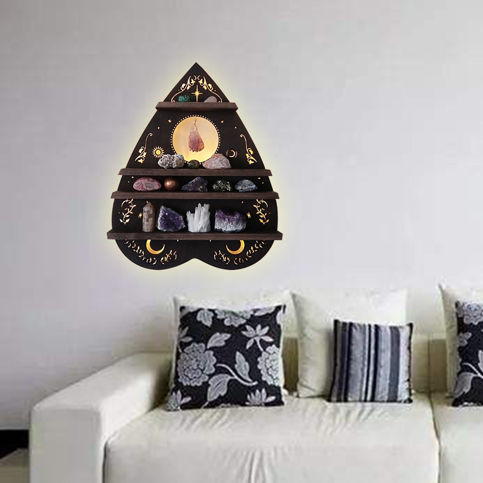 Planchette-shaped celestial shelf mounted above a white sofa with patterned cushions, showing its tiered crystal display in a living-room arrangement.
