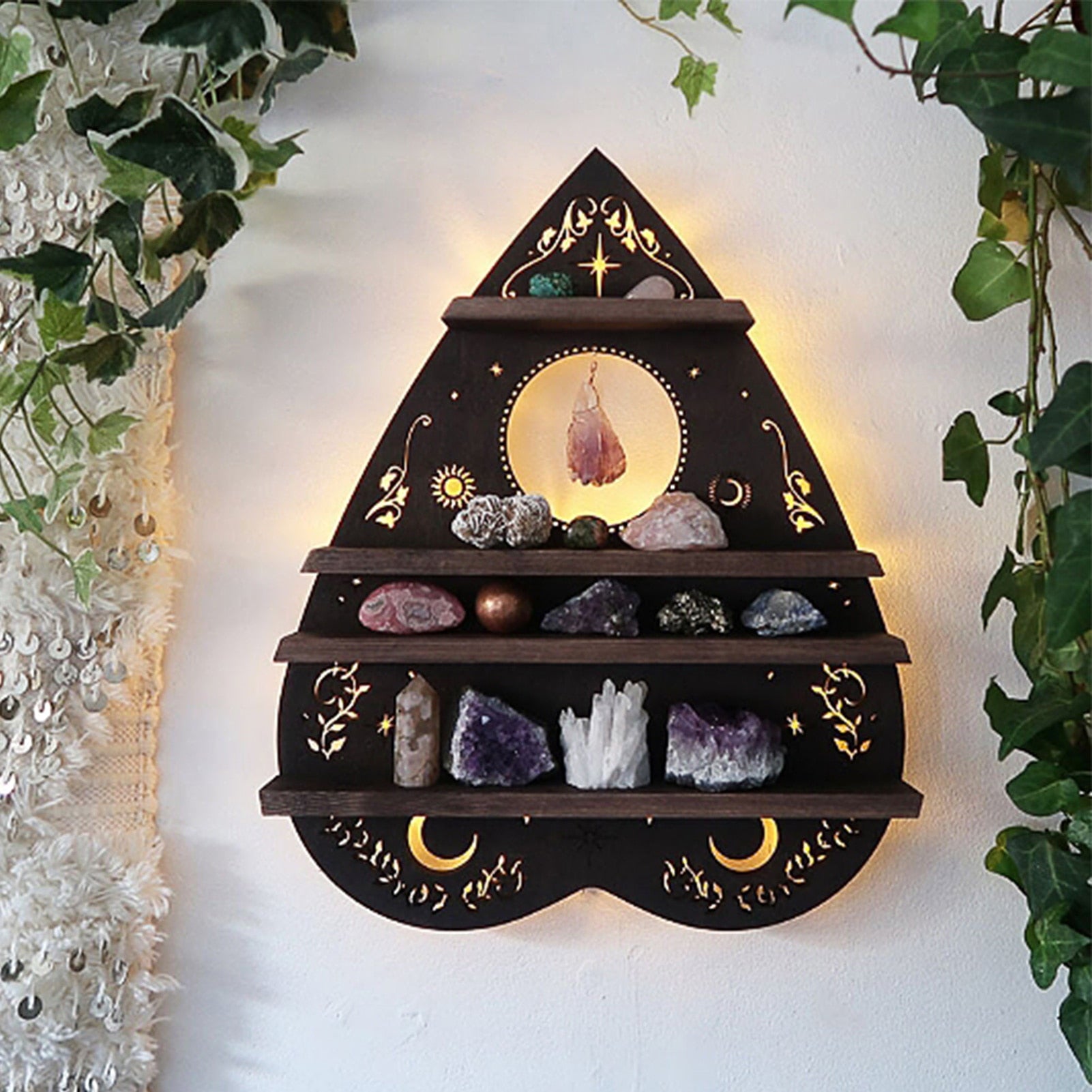 Close wall display of the dark wooden planchette shelf framed by pale leaves, showing golden moon and vine motifs and four ledges of stones.