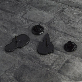 Reverse sides of the potion and skull-candle pins on gray fabric, with their single posts and separate black butterfly-clutch backs.