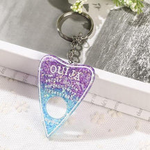 Glitter-filled planchette keychain fading from purple to bright blue, with white lettering and a round opening near the pointed tip, photographed beside an open book.