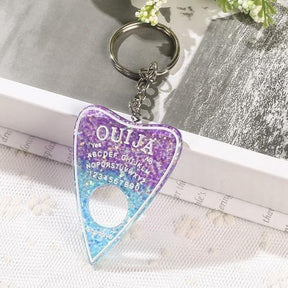 Glitter-filled planchette keychain fading from purple to bright blue, with white lettering and a round opening near the pointed tip, photographed beside an open book.