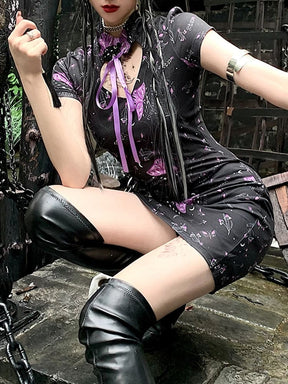 Angled seated view of the black moth-print dress with short sleeves, a high collar and a purple ribbon above the chest opening.