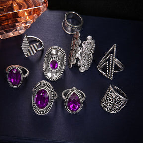 Nine-piece silver-tone ring set with purple jewel-like accents arranged on a dark surface beside a decorative bottle.