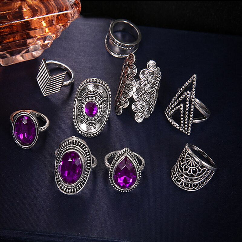 Nine-piece silver-tone ring set with purple jewel-like accents arranged on a dark surface beside a decorative bottle.