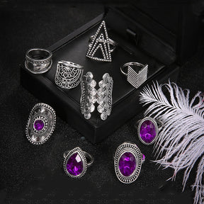 Nine-piece silver-tone ring set with purple jewel-like accents arranged around a dark jewelry box.