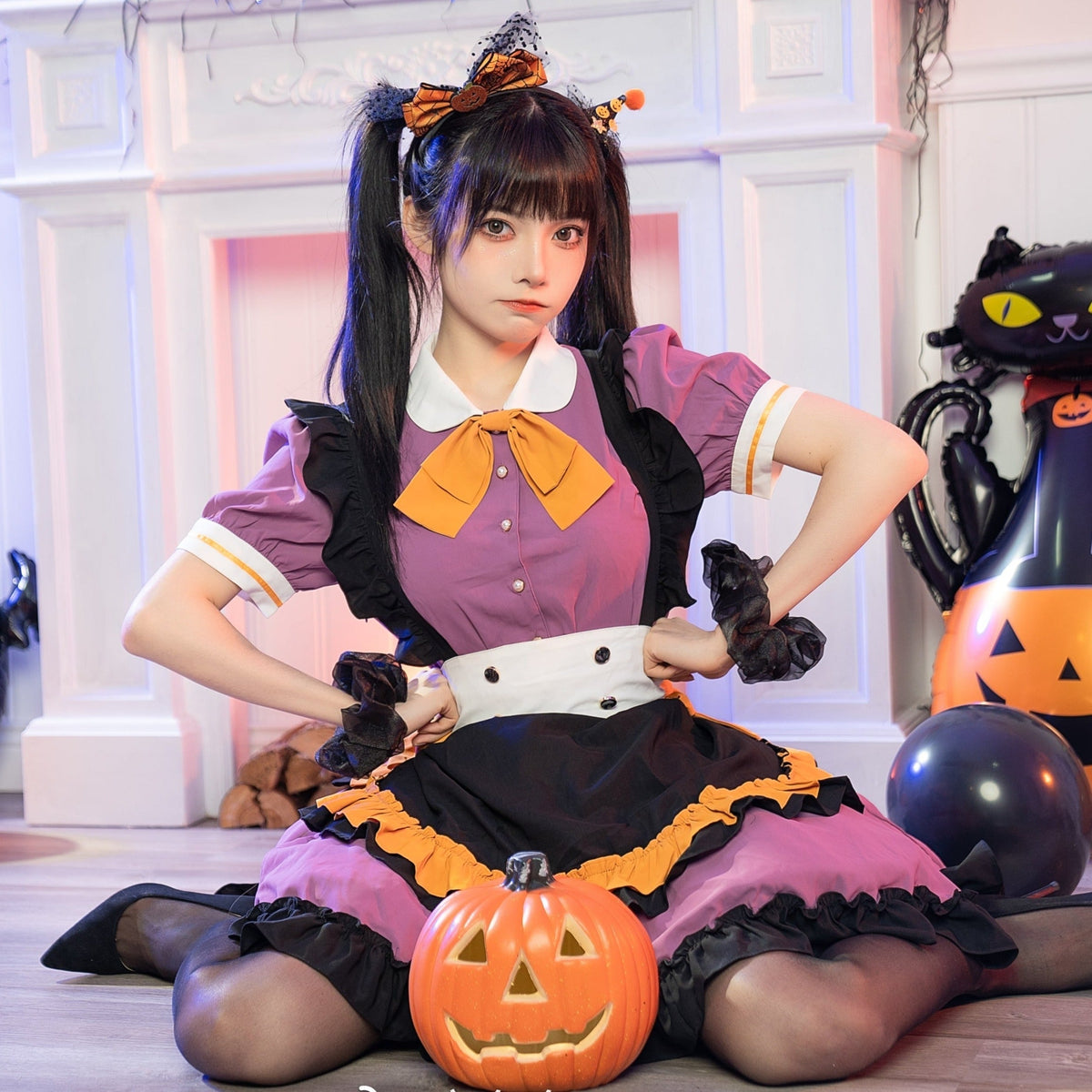 Purple Halloween ruffle dress with an orange neck bow, white collar and black apron-style front. A seated model holds a pumpkin bucket beside black cat decorations.