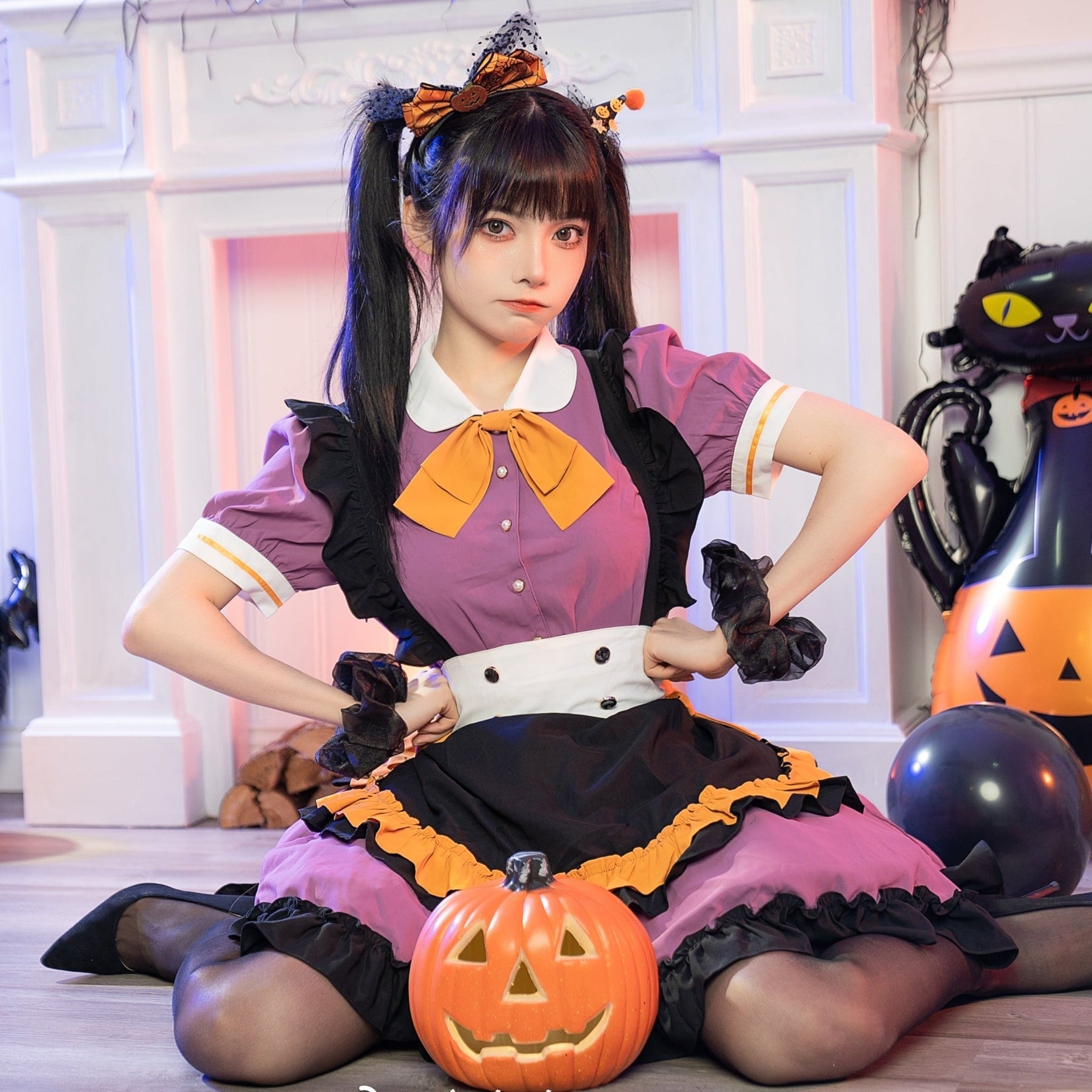 Purple Halloween ruffle dress with an orange neck bow, white collar and black apron-style front. A seated model holds a pumpkin bucket beside black cat decorations.