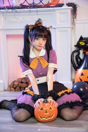 Seated model resting both hands on a pumpkin bucket while wearing the purple and black ruffle dress, showing the white collar and waist panel.