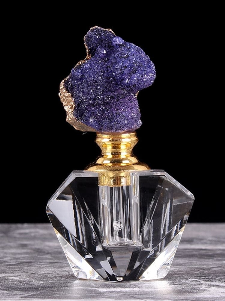 Purple crystal cluster stopper above a clear angular perfume bottle on a stone-like surface against black.