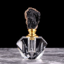 Black crystal cluster stopper with metallic-tone edging on a clear faceted perfume bottle, displayed on a pale textured surface.