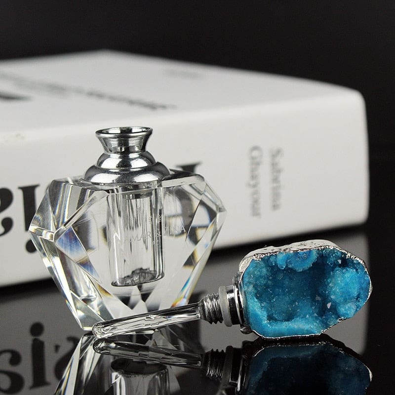 Open faceted perfume bottle beside a blue crystal stopper and its long slim dauber, arranged on a reflective surface near a book.