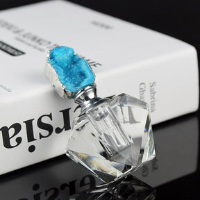 Blue crystal-topped perfume bottle lying at an angle on a reflective surface, showing the clear faceted body and metallic-tone neck.
