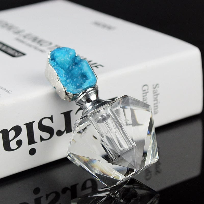 Blue crystal-topped perfume bottle lying at an angle on a reflective surface, showing the clear faceted body and metallic-tone neck.