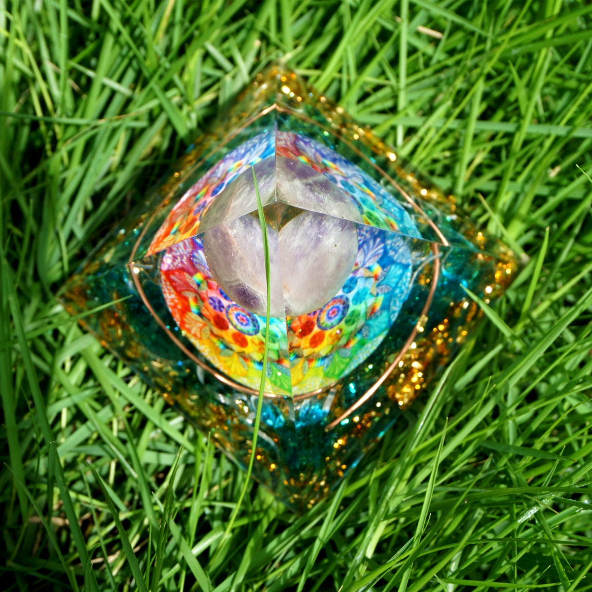 Rainbow mandala orgone pyramid with a pale purple sphere, blue stone details and gold-tone flecks. Viewed from above on grass, showing the multicoloured mandala around the sphere.