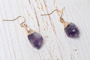 Amethyst hook earrings on pale wood, showing irregular purple stones and gold-tone caps.