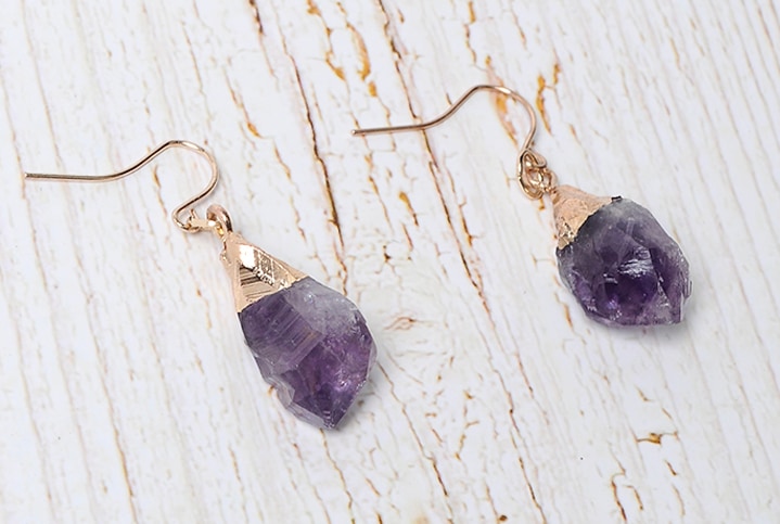 Amethyst hook earrings on pale wood, showing irregular purple stones and gold-tone caps.