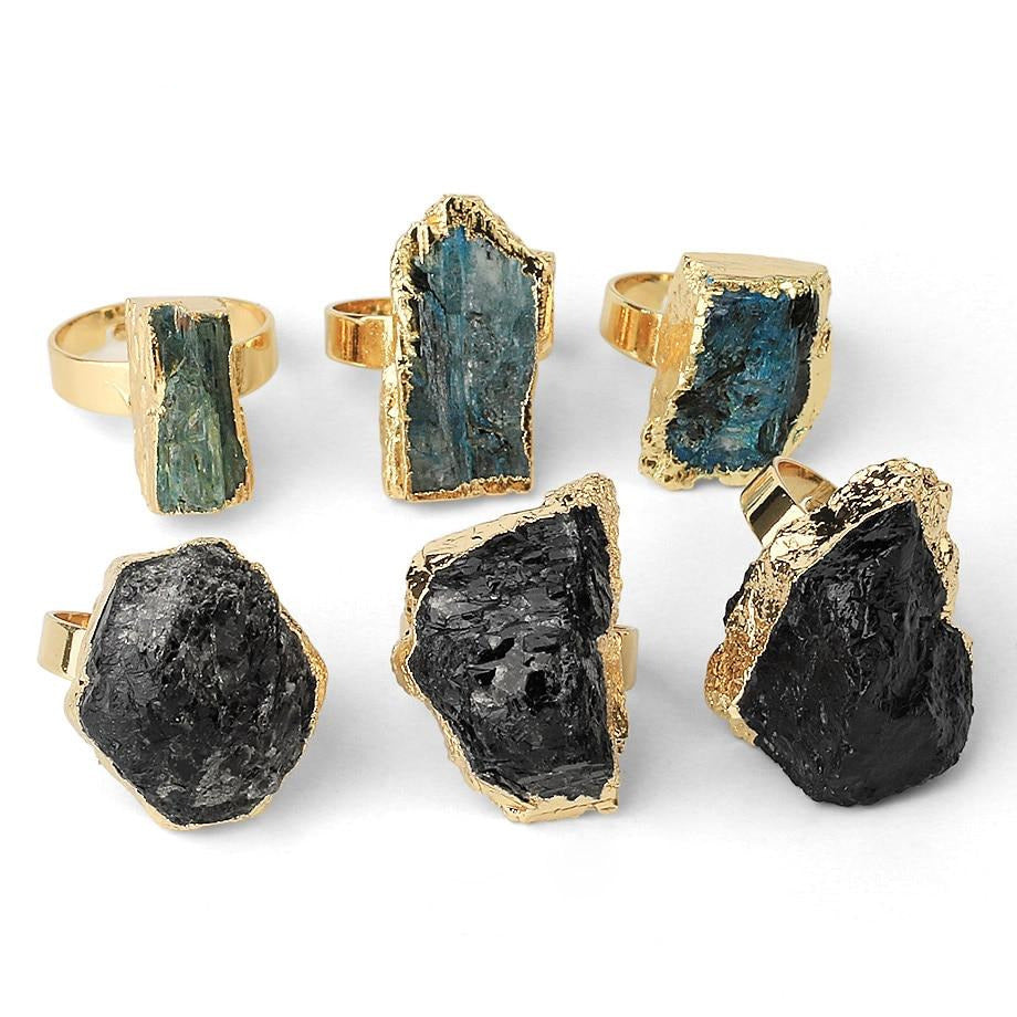 An assortment of raw-look stone rings with gold-tone edges, showing blue, black and other stone examples together.