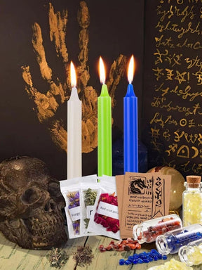White, green and blue slender ritual candles arranged on an altar with a skull and open book, showing the candles among display props.