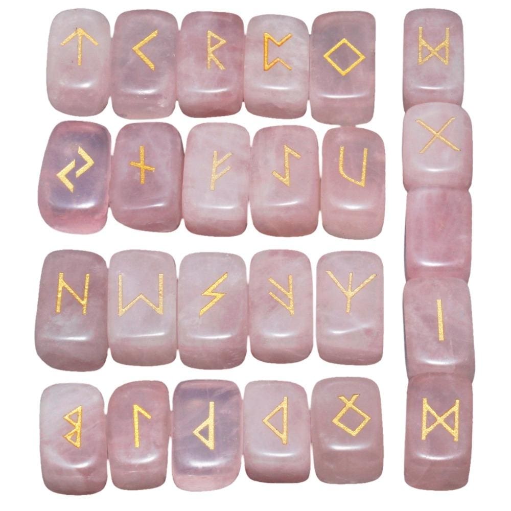 Rose quartz rune stone set laid out in rows, showing individual gold-coloured characters and one unmarked stone against pale pink surfaces.
