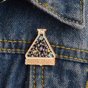 Science, Bitch enamel pin fastened beside a denim seam, with a gold-tone flask outline and small cosmic illustrations inside.