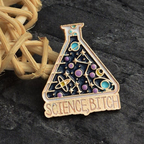 Cosmic science flask pin on gray fabric near a woven ornament, showing turquoise planets, purple circles and the slogan below the flask.
