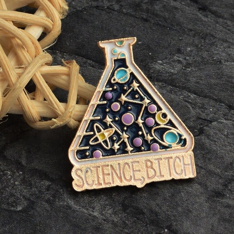 Cosmic science flask pin on gray fabric near a woven ornament, showing turquoise planets, purple circles and the slogan below the flask.