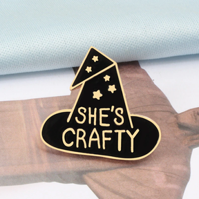 Black She's Crafty enamel pin with a folded hat point, gold-tone stars and lettering.