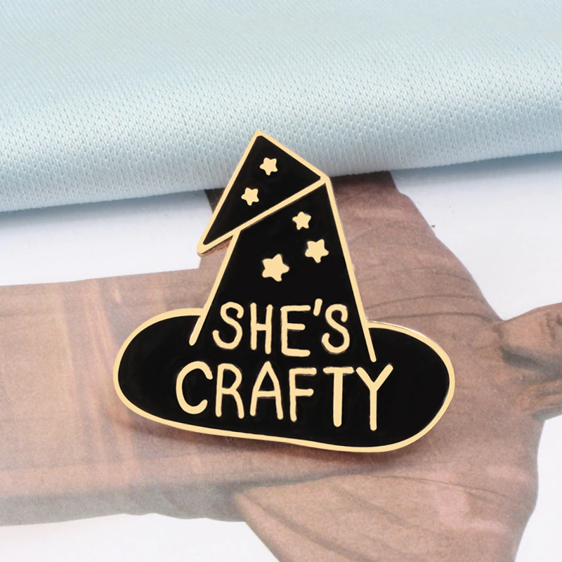 Black She's Crafty enamel pin with a folded hat point, gold-tone stars and lettering.