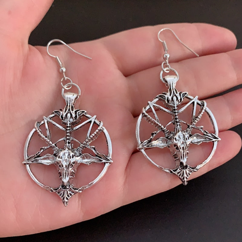 Pair of silver-tone Baphomet pentagram earrings resting on a palm. The open circular frames surround horned-goat faces and branching details.